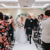 o	Bride and groom walking down the aisle holding hands with white carpet and floral archway, as guests applaud after Scottish last minute wedding