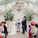 Bride and groom standing with the officiant during a wedding ceremony inside the Pavilion at Cornhill Castle in Biggar, Scotland, decorated with cascading greenery, chandeliers, and large white floral arrangements