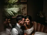 A bride and groom sit together on a couch in a dimly lit room, sharing a quiet moment of intimacy.
