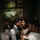 A bride and groom sit together on a couch in a dimly lit room, sharing a quiet moment of intimacy.