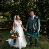 A couple dressed in formal wedding attire laugh beneath shaded trees on the grounds of Bowfield Hotel & Spa, Renfrewshire.