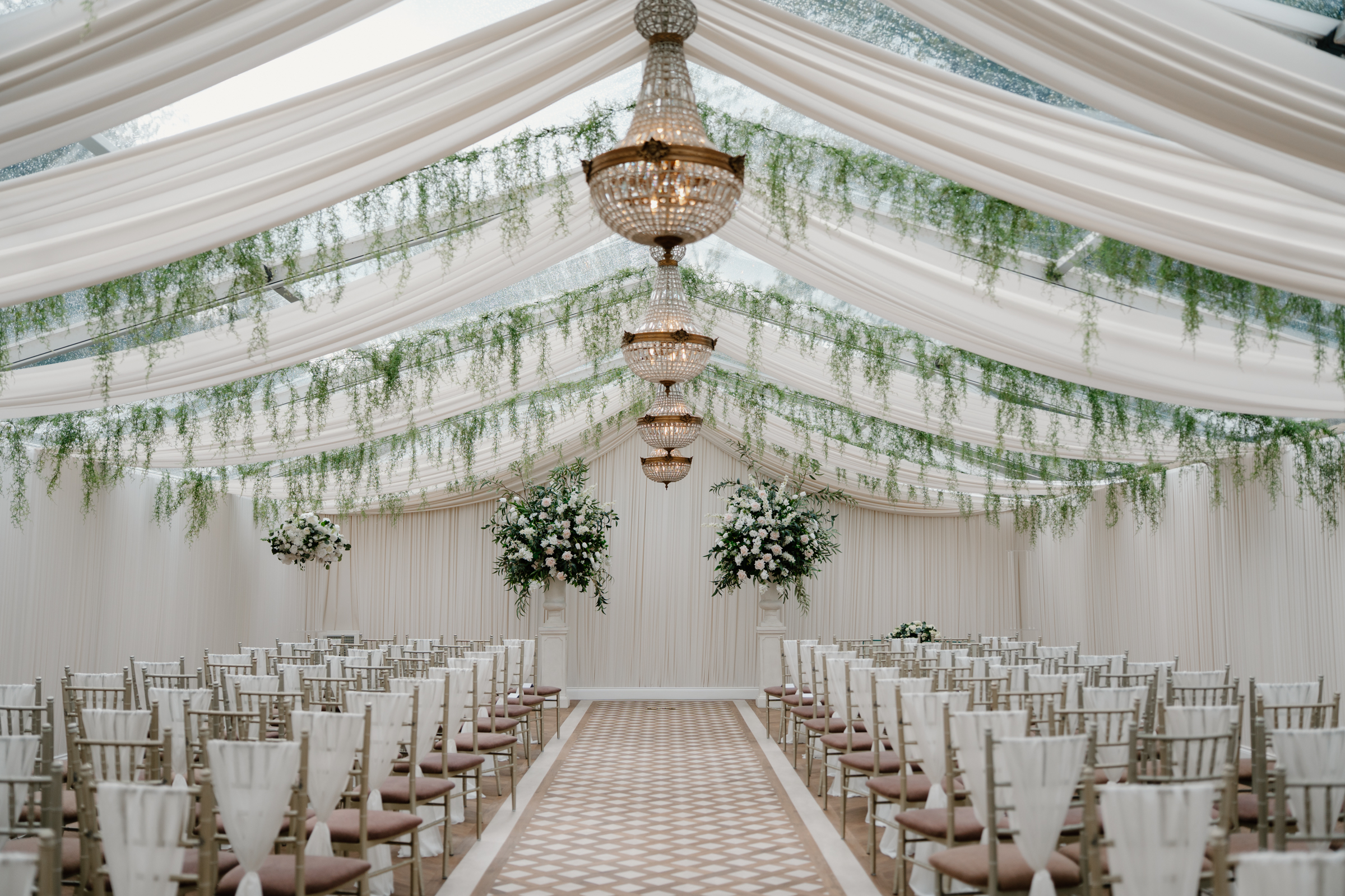 Elegant wedding ceremony setup inside the Pavilion at Cornhill Castle in Biggar, Scotland, with cascading greenery, a chandelier, white floral arrangements, and rows of chairs lining a patterned aisle.