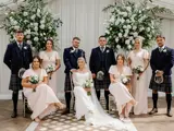 A wedding party smiles for a photo in front of a white wedding arch adorned with flowers and greenery inside the Pavilion at Cornhill Castle.