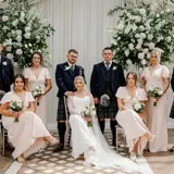 A wedding party smiles for a photo in front of a white wedding arch adorned with flowers and greenery inside the Pavilion at Cornhill Castle.