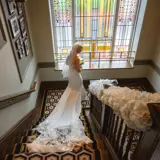 A bride descending the staircase at Boclair House in Bearsden, a Glasgow wedding venue, with her long lace train flowing behind her beside floral décor and stained-glass windows.