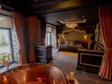 Luxurious honeymoon suite interior at The Garden House, Redhurst Hotel, with a grand bed, copper bathtub by large windows overlooking a garden, and elegant dark décor.