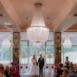 A couple standing at altar during elegant indoor ceremony at Boclair House, Bearsden, a Glasgow wedding venue with grand chandeliers, floral décor and guests either side of the aisle.