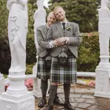 Two grooms wearing matching kilts standing in a stone pergola in a garden