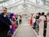 Wedding guests standing along the aisle inside the Pavilion at Cornhill Castle in Biggar, Scotland, decorated with hanging greenery and chandeliers, awaiting the ceremony procession