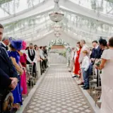 Wedding guests standing along the aisle inside the Pavilion at Cornhill Castle in Biggar, Scotland, decorated with hanging greenery and chandeliers, awaiting the ceremony procession