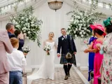 Happy couple walk down the aisle at Cornhill Castle in Biggar, Scotland, greeted by guests and surrounded by white floral arrangements