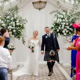 Happy couple walk down the aisle at Cornhill Castle in Biggar, Scotland, greeted by guests and surrounded by white floral arrangements