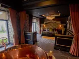 Luxurious honeymoon suite interior at The Garden House, Redhurst Hotel, with a grand bed, copper bathtub by large windows overlooking a garden, and elegant dark décor.